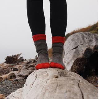Person in black leggings and hand-knit gray socks with red striped cuffs standing on a driftwood log at a rocky beach