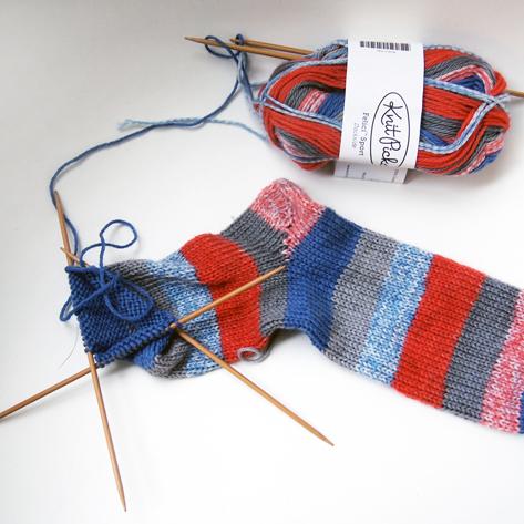 In-progress striped sock on wooden double-pointed needles in red, blue, gray, and pink with matching Knitpicks yarn skein