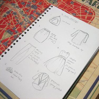 Spiral notebook with hand-drawn fashion sketches of tops, skirts, and jacket designs resting on a Paris map