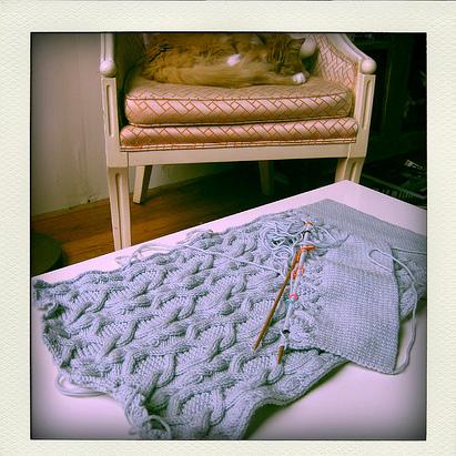 Blue cabled knit vest in progress with wooden needles on white surface, cat resting in cream armchair in background