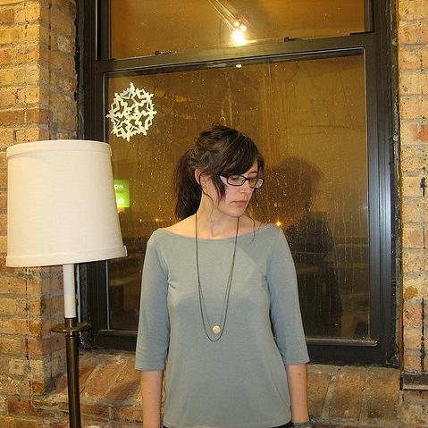 Person wearing gray three-quarter sleeve top with boat neckline and pendant necklace against a brick wall and window