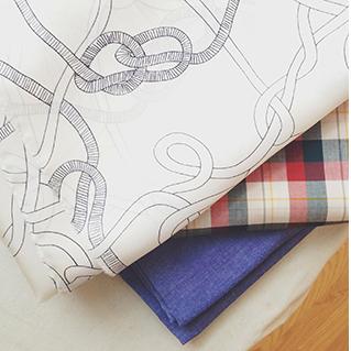 Stack of fabrics: cream silk with black rope print, red and blue plaid cotton shirting, and blue chambray