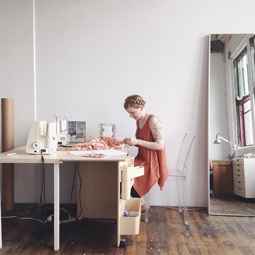 Person in rust sleeveless top working at sewing table with serger in bright studio workspace with floor length mirror