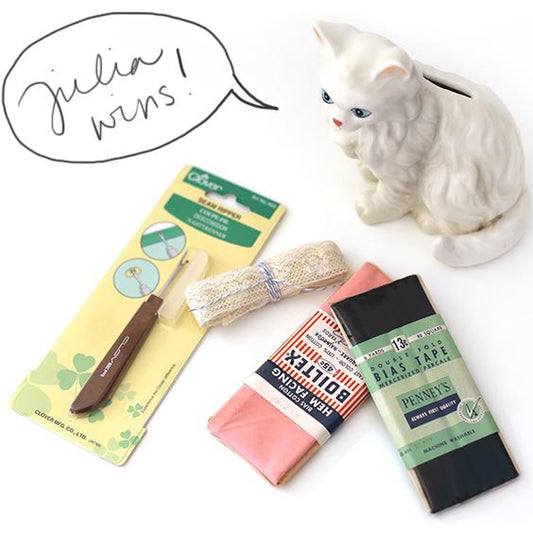 Giveaway flat lay with a white ceramic cat figurine, Clover seam ripper, lace trim, and vintage bias tape packages
