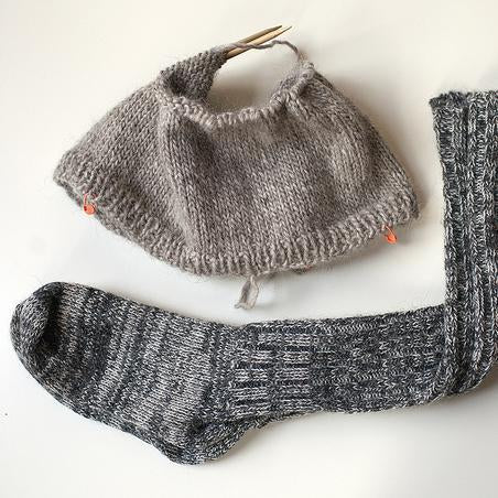 Gray stockinette knit sweater with ribbed trim, charcoal cabled knit socks, and partial knitted piece on wooden needles