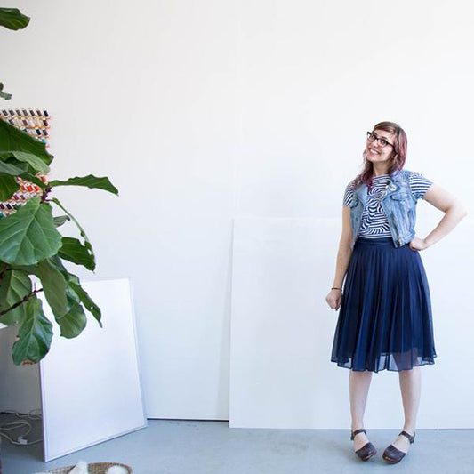 Person in striped top, navy pleated midi skirt, and denim vest, standing against a white wall with a potted fiddle leaf fig