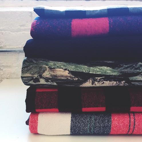 Stack of folded fabrics in navy and pink buffalo plaid, green camo, and red and cream plaid against white brick wall