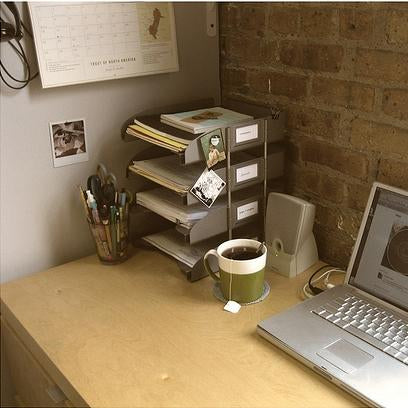 Sewing studio desk with paper organizer, pencil holder, coffee mug, and laptop against exposed brick wall