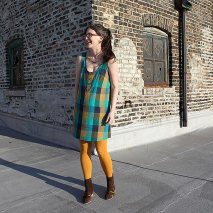 Teal and mustard plaid sleeveless shift dress with button placket, worn with golden tights and brown boots against brick wall