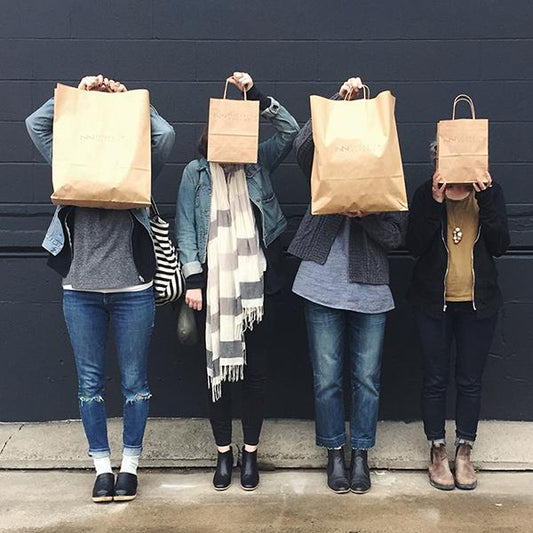 Four people holding paper bags over their faces in casual layered outfits with jeans and boots against a dark wall