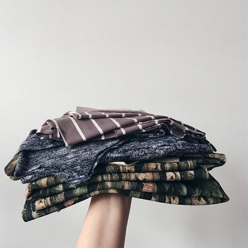 Hand holding a stack of folded knit fabrics in navy marl, brown stripe, and camo print on white background