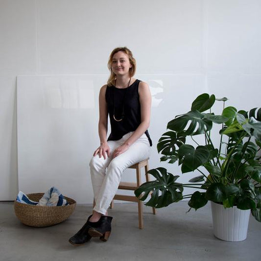 Person in black sleeveless top and white pants seated on a wooden stool beside a monstera plant and woven basket