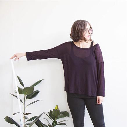 Person wearing a dark purple long-sleeved Hemlock Tee and black pants, posing next to a potted plant