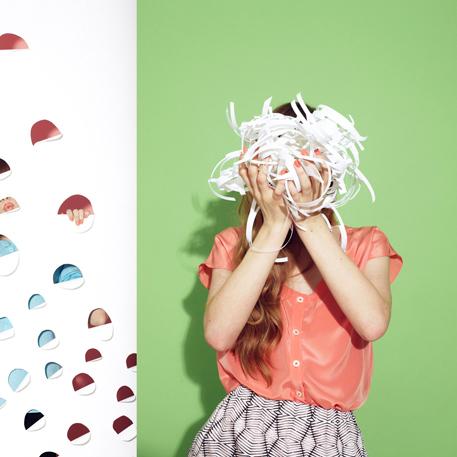 Person in coral top and geometric skirt holding shredded paper over face against green wall with colorful half-circle swatches