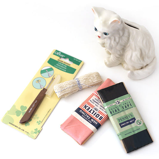 Flat lay of sewing notions including a white ceramic cat figurine, Clover seam ripper, lace trim, and vintage bias tape packages