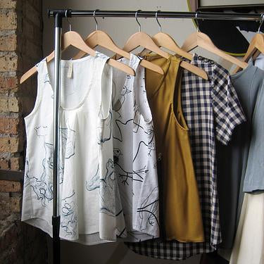 Handmade tops on wooden hangers including white printed tanks, mustard top, gingham shirt, and gray top against brick wall