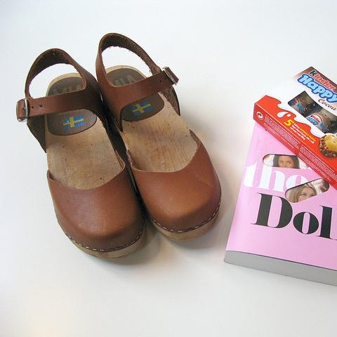 Pair of brown leather Swedish clogs with wooden soles and ankle straps next to Valley of the Dolls book and Kinder Chocolate