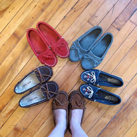 Six moccasins arranged on wood floor in red, gray, dark brown, navy with flag embroidery, and tan suede styles
