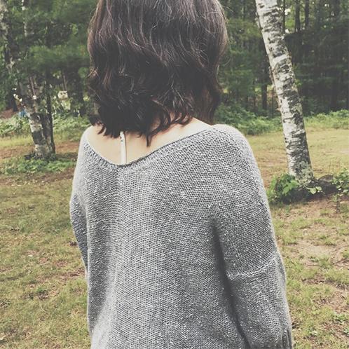 Person wearing gray textured knit Lucinda sweater, photographed from behind in a forest clearing with birch trees