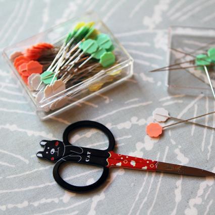 Black cat-shaped embroidery scissors with red patterned handle next to clear container of colorful sewing pins on gray fabric
