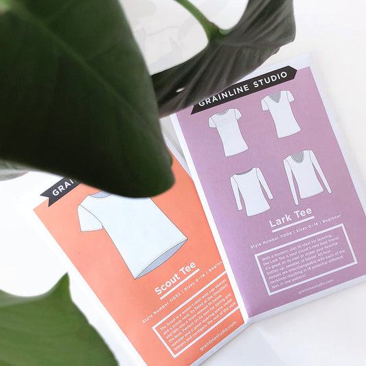 Grainline Studio Scout Tee pattern envelope in coral and Lark Tee envelope in mauve, fanned out under plant leaves