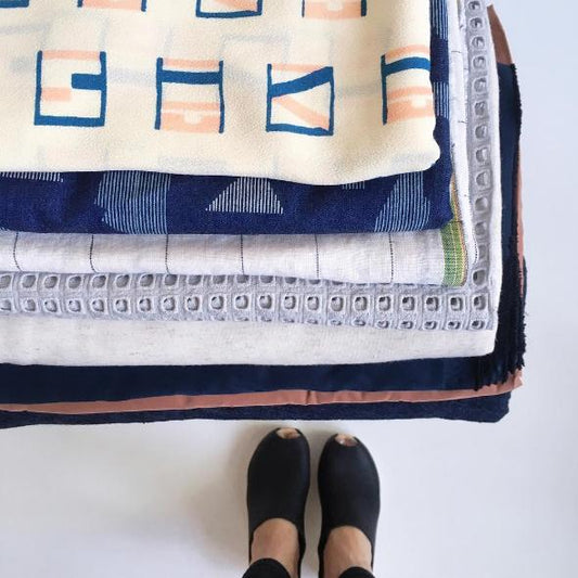Stack of folded fabrics in geometric print, navy stripe, gray eyelet, white linen, and navy velvet with feet in black shoes