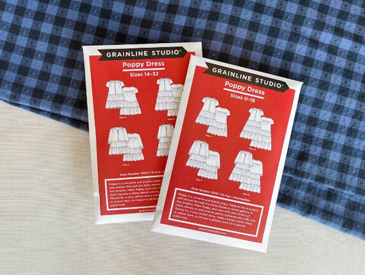 Two red Grainline Poppy Dress sewing pattern envelopes with line drawings, sizes 0-18 and 14-32, on blue gingham fabric