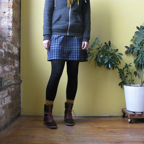 Person wearing gray zip-up hoodie, blue plaid skirt, black tights, mustard socks, and brown lace-up boots against yellow wall