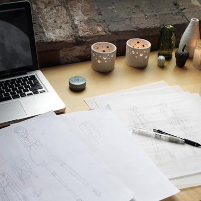 Wooden desk with fashion sketches, a marker, laptop, ceramic candle holders, and bottles against an exposed brick wall