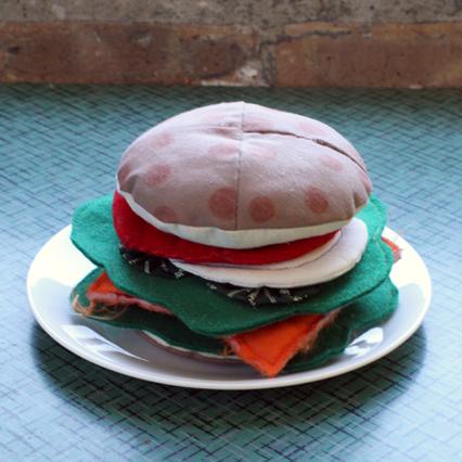 Handmade felt burger with tomato, onion, lettuce, and cheese on a white plate against a turquoise tile background