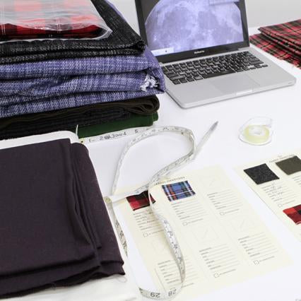 Folded fabric samples in plaid, wool, and jersey with fabric swatch cards, measuring tape, and an open laptop on a white table