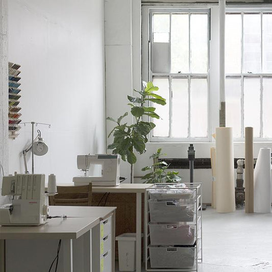 Grainline Studio workspace with sewing machines, serger, storage drawers, fiddle leaf fig, thread rack, and large windows