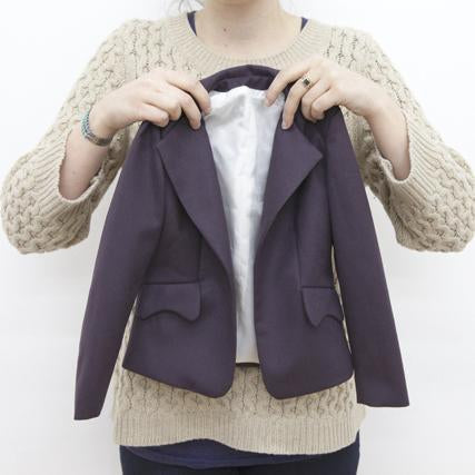 Person holding a mini purple blazer with notch lapels and white lining in front of a cream cable knit sweater