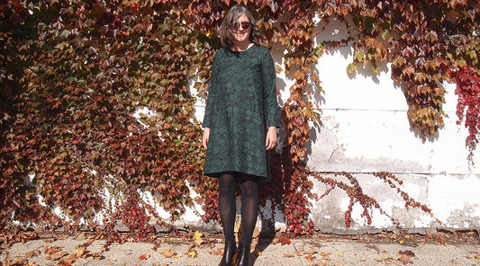 Farrow Dress in green and black print with long sleeves and black tights, standing in front of an autumn ivy-covered wall