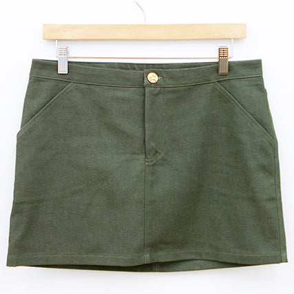 Moss Skirt in olive green denim with angled front pockets and gold button closure on a wooden hanger