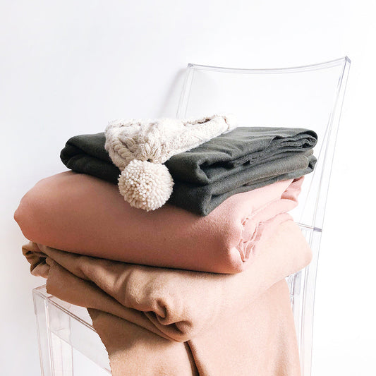Folded wool coating fabrics in camel, blush, and olive stacked on a clear acrylic chair with a cream pom-pom hat