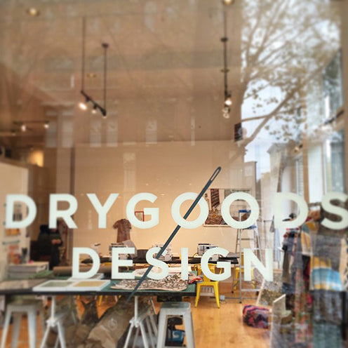 Drygoods Design storefront window with white lettering and interior showing sewing supplies, pendant lights, and wood furniture