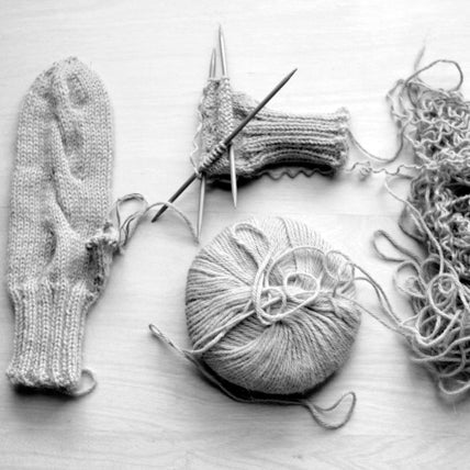 Flat lay of a knit mitten, mitten in progress on circular needles, ball of yarn, and tangled yarn scraps on white surface