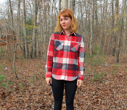 Person wearing red, white, and navy plaid Archer Button Up shirt in cotton flannel, standing outdoors in a forest setting