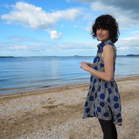 Woman wearing navy large polka dot Alder Shirtdress with funnel neck on a beach with blue water and sky in background