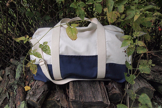 Portside Travel Set duffle in natural canvas with navy colorblock and webbing straps, resting on a log surrounded by foliage