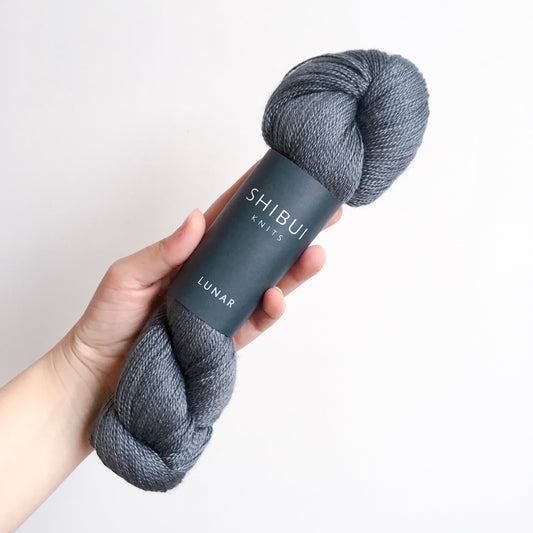 Hand holding a skein of Shibui Knits Lunar yarn in dark slate blue with branded paper band