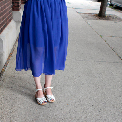 Cobalt blue chiffon midi skirt with gathered waist styled with white strappy sandals on a sidewalk