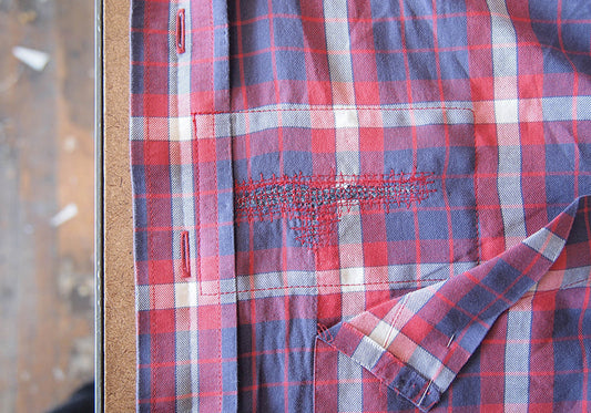 Plaid brushed cotton shirt with visible mending stitches beneath pocket, shown with pocket partially detached and folded back