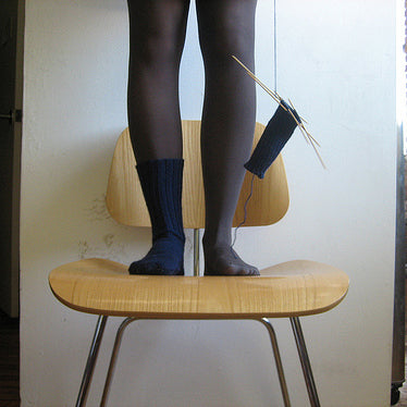 Person standing on wooden chair wearing black ribbed knit socks with navy cuffs, holding knitting needles and yarn