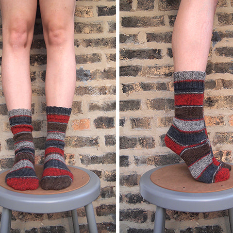Front and side views of handknit striped socks in gray, red, and brown, standing on a stool against a brick wall