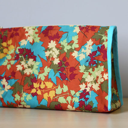 Colorful autumn leaf print pouch in red, orange, yellow, cream, and teal on orange background, bound with teal linen trim