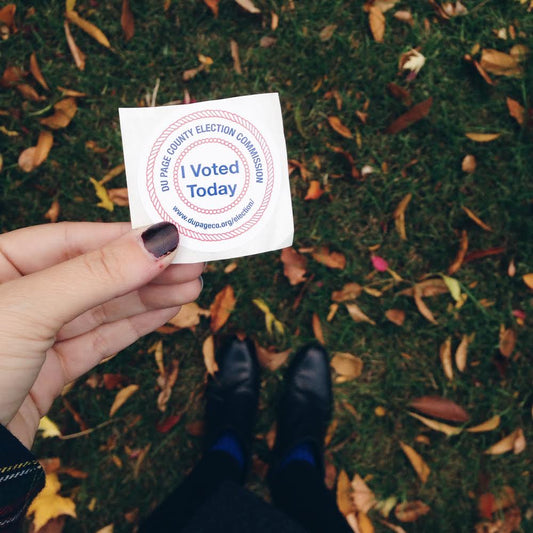 Person holding "I Voted Today" sticker from Du Page County Election Commission over autumn leaves and feet in black shoes