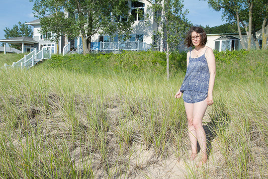 Woman wearing a blue printed Lakeside Pajamas camisole and shorts, barefoot in sand dunes with a beach cottage behind her