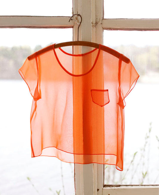 Cropped Scout Tee in orange silk organza with short sleeves and chest pocket, hanging on wooden hanger in front of window